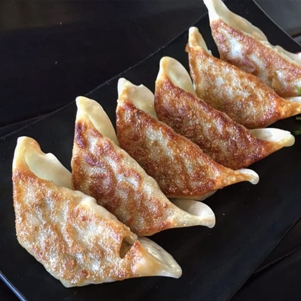 Six pan-fried gyoza with crispy, golden-brown bottoms arranged on a black rectangular plate.